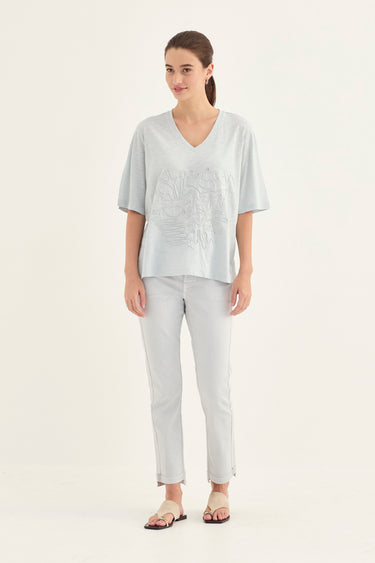 A woman modeling a light gray short-sleeve V-neck top with a tonal embroidered chest design, paired with matching light gray pants by Verge and flat sandals, against a plain white studio background.