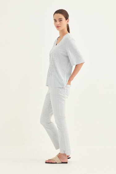 A woman modeling a light gray linen scrub set consisting of a short-sleeve V-neck top and matching straight-leg pants by Verge, paired with beige slide sandals, against a plain white studio background.
