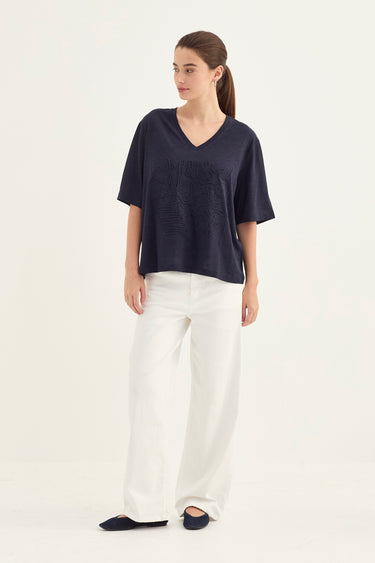 A woman modeling a navy blue short-sleeve V-neck top with textured embroidery on the front, paired with white wide-leg trousers and dark navy flats by Verge, against a plain white studio background.