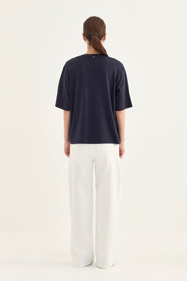 A woman viewed from behind wearing a dark navy blue short-sleeve t-shirt and white wide-leg trousers by Verge, standing against a plain white studio background.