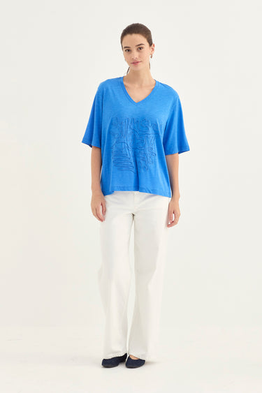 A woman modeling a bright blue short-sleeve V-neck top with a tonal embroidered graphic on the front, paired with white wide-leg pants and dark flats by Verge, against a plain white studio background.