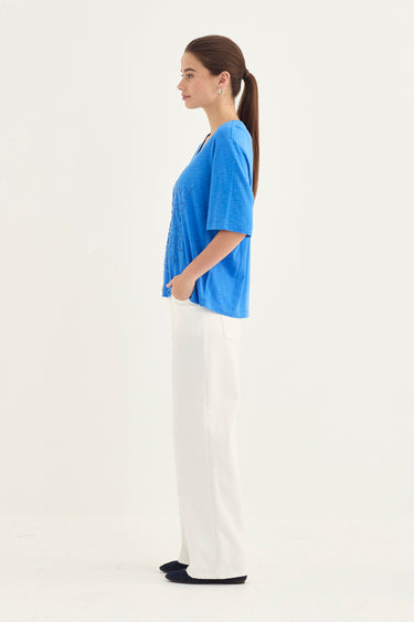A woman shown in profile wearing a bright blue short-sleeve top and white wide-leg pants by Verge against a plain white studio background.