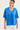 A woman wearing a bright blue short-sleeve V-neck t-shirt with a tonal embroidered abstract pattern on the front, paired with white pants by Verge, against a plain light background.