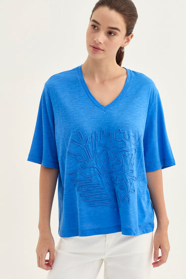 A woman wearing a bright blue short-sleeve V-neck t-shirt with a tonal embroidered abstract pattern on the front, paired with white pants by Verge, against a plain light background.