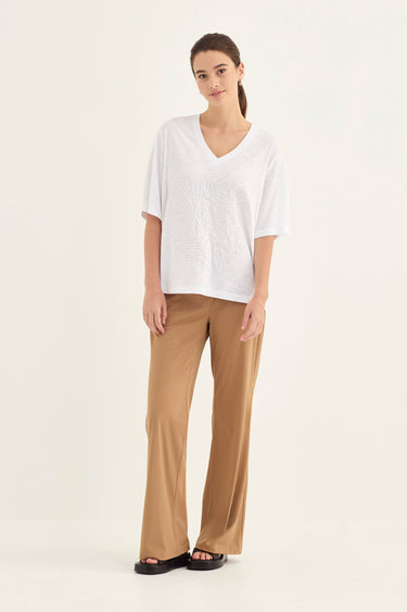 A woman modeling a white short-sleeve V-neck top and tan wide-leg pants by Verge against a plain light background.