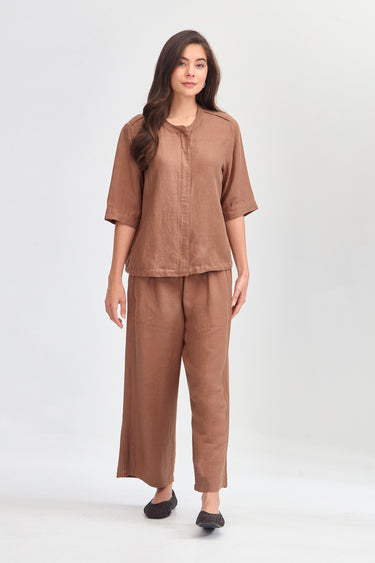 A woman modeling a matching two-piece linen set in a warm brown color, featuring a short-sleeve button-up top and wide-leg trousers by Verge, photographed against a neutral light gray studio background.