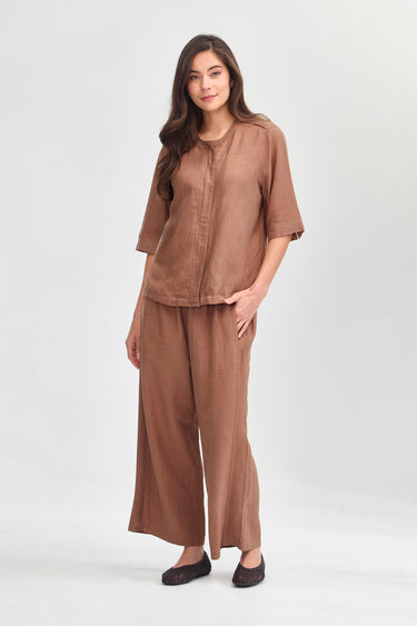 A woman modeling a matching two-piece linen set in a warm brown color, consisting of a short-sleeve button-up top and wide-leg pants by Verge, against a plain light gray background.
