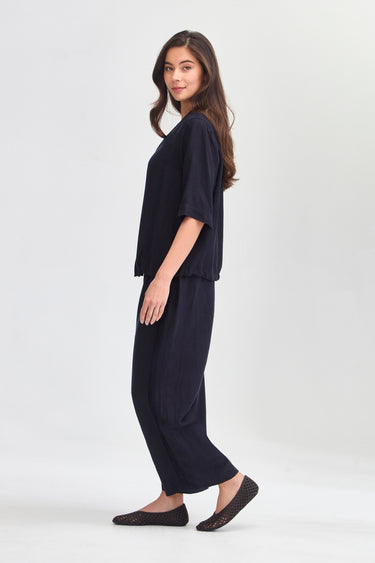 A woman modeling a dark navy blue two-piece lounge set consisting of a short-sleeve top and matching wide-leg pants by Verge, paired with dark slip-on flats, against a light gray studio background.