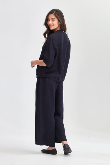 A woman modeling a dark navy blue linen two-piece set consisting of a cropped long-sleeve top and matching wide-leg trousers by Verge, paired with black flats, against a plain white studio background.