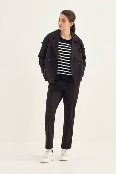 A woman modeling a black double-breasted blazer, black trousers by Verge, a black and white striped t-shirt, and white sneakers against a plain light gray studio background.