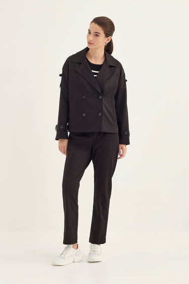 A woman modeling a black double-breasted blazer with shoulder tab details, paired with black trousers by Verge and white sneakers, against a plain light background.