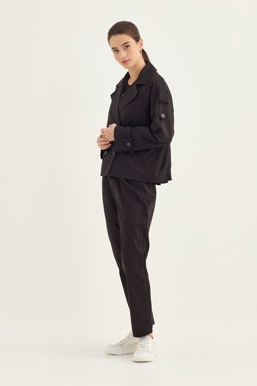 A woman modeling a black utility jacket and matching black trousers by Verge with white sneakers, standing against a plain white studio background.