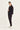 A woman modeling a black utility jacket and matching black trousers by Verge with white sneakers, standing against a plain white studio background.