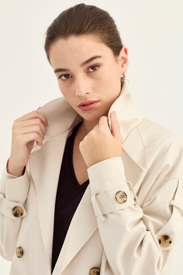 A woman wearing a light beige double-breasted trench coat over a black top by Verge, posing against a plain white studio background.
