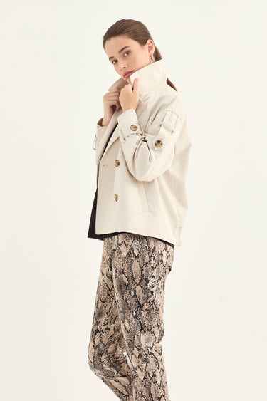 A woman modeling a light beige double-breasted jacket with gold buttons, worn over a black top and paired with beige and black snakeskin print trousers by Verge, against a plain off-white background.