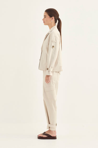 A woman in side profile wearing a light beige tailored jacket and matching trousers by Verge with dark brown flip-flop sandals, photographed against a plain off-white studio background.