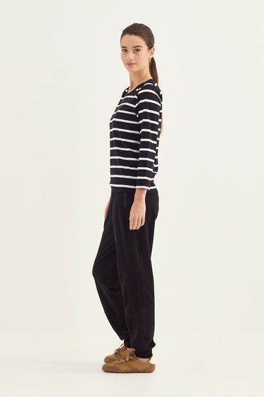 A woman stands in profile wearing a black and white horizontally striped long-sleeve top, black wide-leg pants, and brown slip-on shoes by Verge against a plain light background.