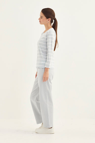 A woman in a side profile wearing a light gray and white striped long-sleeve top, matching wide-leg trousers, and white sneakers by Verge, set against a plain white studio background.