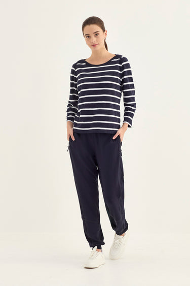 A woman modeling a navy blue and white horizontally striped long-sleeve top with dark navy jogger pants by Verge and white sneakers, standing against a plain off-white studio background.