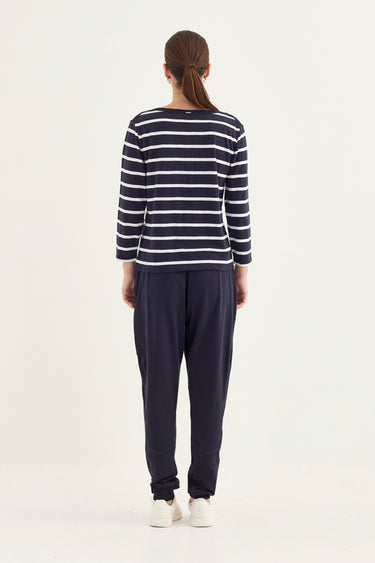 A woman viewed from behind wearing a navy and white horizontally striped long-sleeve top, dark navy pants by Verge, and white sneakers, against a plain light background.