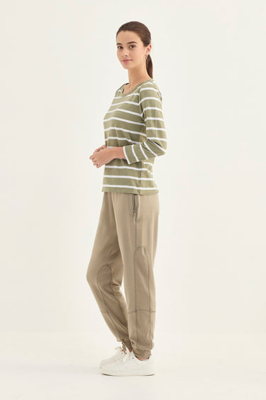 A woman modeling a long-sleeve crewneck top with horizontal olive green and white stripes, paired with beige jogger pants and white sneakers by Verge, set against a plain white studio background.