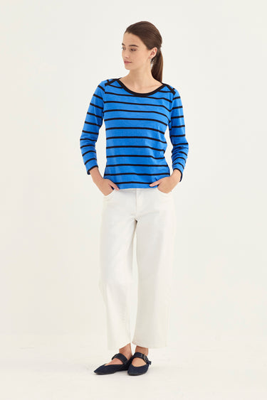 A woman modeling a blue and black striped long-sleeve top with white pants and black flats by Verge, standing against a plain white studio background.