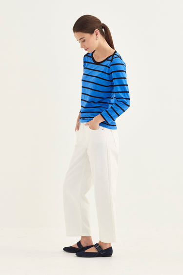 A woman modeling a blue and black striped long-sleeve top with white cropped pants and black flat shoes by Verge, standing against a plain white studio background.