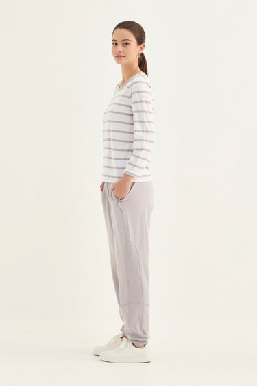 A woman stands in profile wearing a white long-sleeve top with thin gray horizontal stripes, light gray jogger pants, and white sneakers by Verge against a plain white studio background.