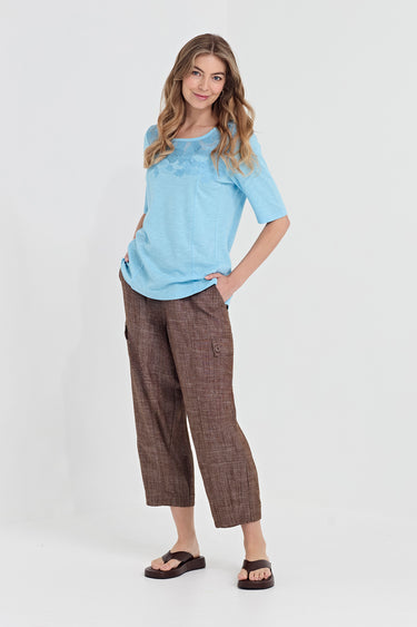 A woman modeling a light blue short-sleeve t-shirt and brown cropped trousers with a subtle plaid pattern by Verge, paired with dark brown slide sandals, against a plain light gray studio background.