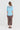A woman viewed from the back wearing a light blue short-sleeve top with a sheer yoke and brown cropped trousers by Verge, standing against a plain light gray studio background.