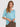 A woman modeling a light blue short-sleeve top with a sheer floral lace overlay on the chest, paired with brown textured pants by Verge, against a solid light blue background.