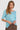 A woman modeling a light blue short-sleeve top with a sheer floral lace overlay on the chest, paired with brown textured pants by Verge, against a solid light blue background.