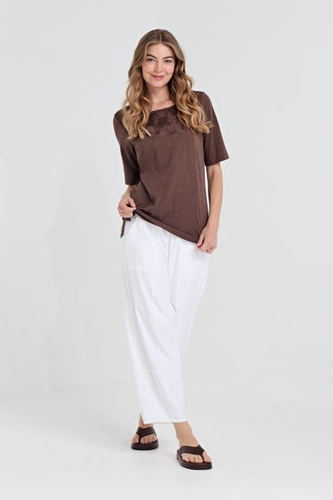A woman modeling a brown short-sleeve top and white wide-leg pants by Verge against a plain light gray studio background.