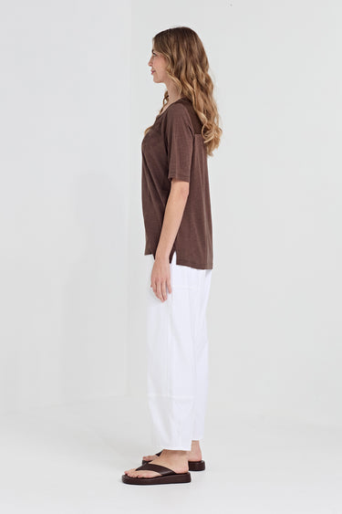 A woman shown in profile wearing a brown short-sleeve top, white pants by Verge, and dark brown sandals against a plain light gray studio background.