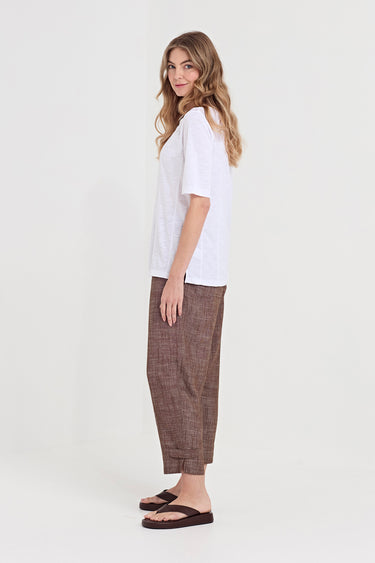 A woman modeling a white short-sleeve top and brown textured cropped pants by Verge with dark brown sandals, standing against a plain light gray studio background.