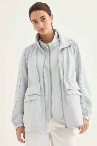 A woman wearing a light gray lightweight zip-up jacket with a stand-up collar and front flap pockets, paired with white pants by Verge, against a plain off-white studio background.