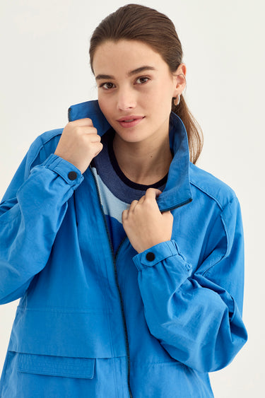 A woman wearing a bright blue utility-style jacket by Verge with a high collar, shown from the chest up against a plain light background.