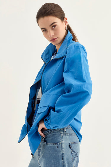 A woman wearing a bright blue lightweight jacket and light-wash blue jeans by Verge, photographed against a plain light background.