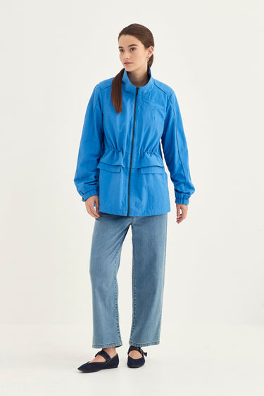 A woman modeling a bright blue lightweight jacket with a high collar, front zipper, and drawstring waist, paired with light blue jeans by Verge and dark flats, against a plain light gray studio background.
