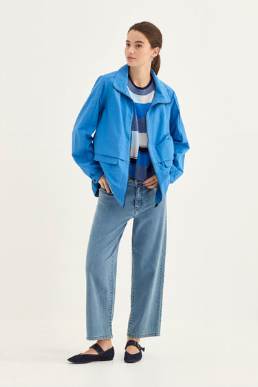 A woman modeling a bright blue lightweight jacket over a striped blue and white top, paired with light-wash wide-leg jeans by Verge and black flats, against a plain off-white studio background.