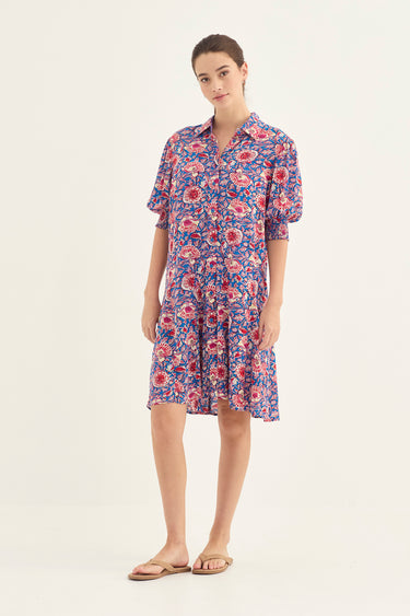 A woman modeling a short-sleeve button-front dress by Verge with a dense floral print in blue, pink, and red, paired with tan flat sandals, against a plain light background.