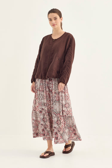 A woman modeling a dark brown long-sleeve top paired with a high-waisted, pleated maxi skirt featuring a dusty rose and beige paisley pattern by Verge, set against a plain off-white studio background.