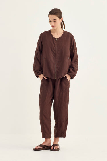 A woman modeling a dark brown two-piece linen set consisting of a cropped zip-up jacket and matching relaxed-fit pants by Verge, paired with brown sandals, against a plain off-white studio background.