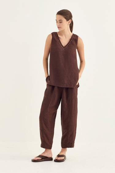 A woman modeling a dark brown sleeveless tank by Verge with a V-neck and decorative trim, paired with matching loose-fit pants and dark sandals, against a plain light background.