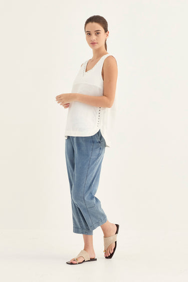 A woman modeling a white sleeveless top with side buttons and light blue cropped pants by Verge, standing against a plain white studio background.