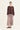 A model stands against a plain off-white studio background wearing a dark brown cropped long-sleeve top and a long pleated maxi skirt with a dusty rose and beige paisley pattern by Verge, paired with dark brown slide sandals.