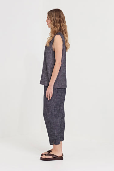 A woman stands in profile wearing a matching sleeveless top and cropped pants set in a dark gray textured fabric by Verge, paired with dark brown flip-flop sandals, against a plain light gray studio background.