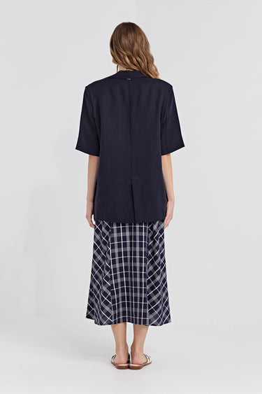 A woman viewed from the back wearing a dark navy short-sleeve button-front top and a navy-and-white plaid dress by Verge, standing against a light gray studio background.