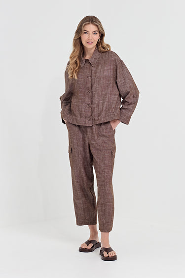 A woman modeling a matching brown plaid button-up shirt and cropped cargo pants set by Verge, paired with dark brown slide sandals, against a plain light gray studio background.