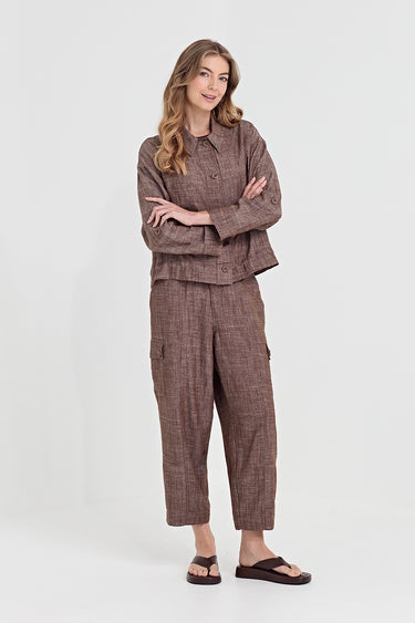 A woman modeling a matching brown plaid two-piece lounge set consisting of a cropped button-up shirt and cropped cargo pants by Verge, paired with dark brown slide sandals, against a plain light gray studio background.